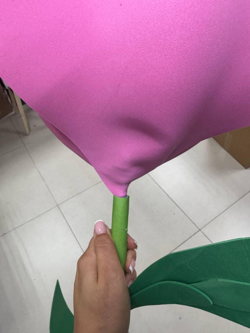 May include: A large pink foam flower head attached to a green cylindrical stem. The flower features a smooth, matte texture and is connected to a green stem that has a single green leaf extending from the side, constructed from foam material.