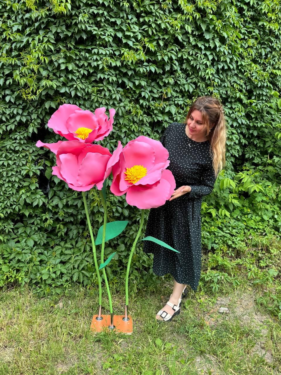 May include: Three large, oversized artificial flowers with vibrant pink petals and textured yellow centers. Each flower is supported by a thin green stem attached to a rectangular wooden base, creating a tall, freestanding decorative display.