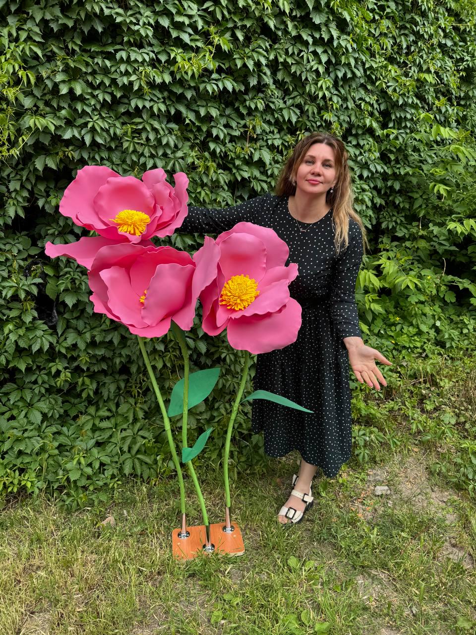 May include: Three oversized artificial flowers with vibrant pink petals and textured yellow centers. Each flower is attached to a thin green stem with a single leaf, all mounted on a rectangular wooden base with metallic hardware connectors.