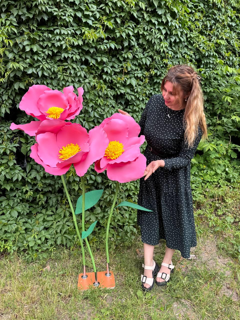 May include: Three large, oversized artificial pink flowers with bright yellow centers, mounted on thin green stems with green leaves, attached to a rectangular wooden base. The flowers feature wide, curved petals and are displayed upright.