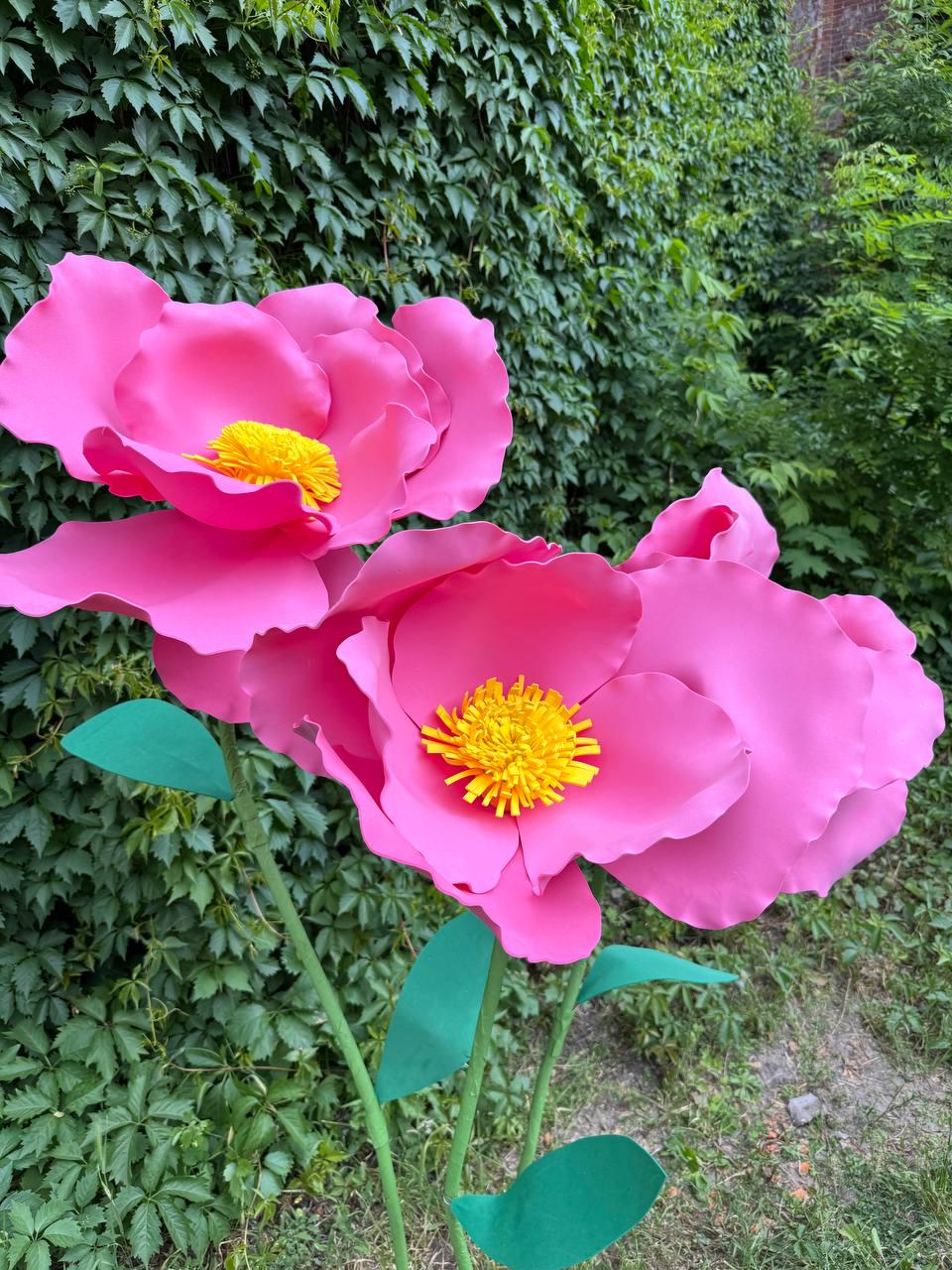 May include: Two large, oversized artificial flowers with vibrant pink, wavy-edged petals and bright yellow, textured centers. Each flower is attached to a thin, green stem featuring several pointed green leaves extending from the stalks.