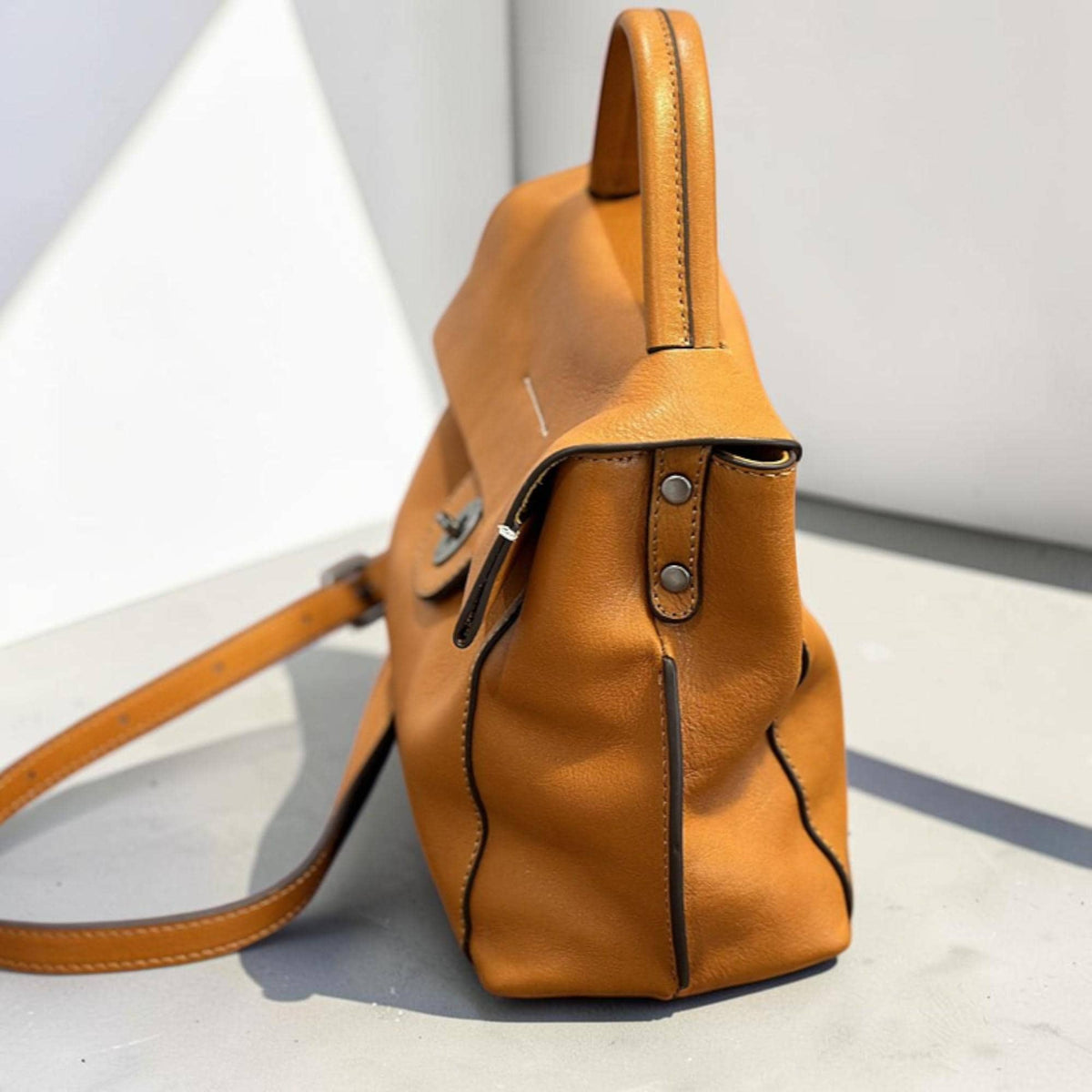 May include: A tan leather satchel bag with a top handle and a long adjustable shoulder strap. The bag has a flap closure with a metal clasp and decorative stitching. The bag is made of a textured leather material.