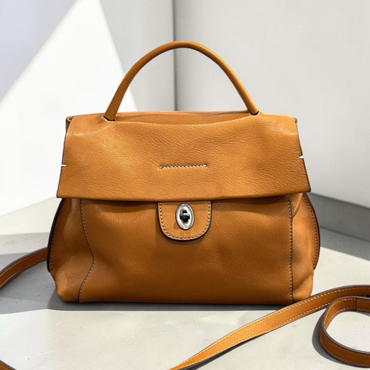 May include: A tan leather handbag with a top handle and a detachable shoulder strap. The bag features a flap closure with a silver-toned clasp. The bag has a structured design and a smooth texture.