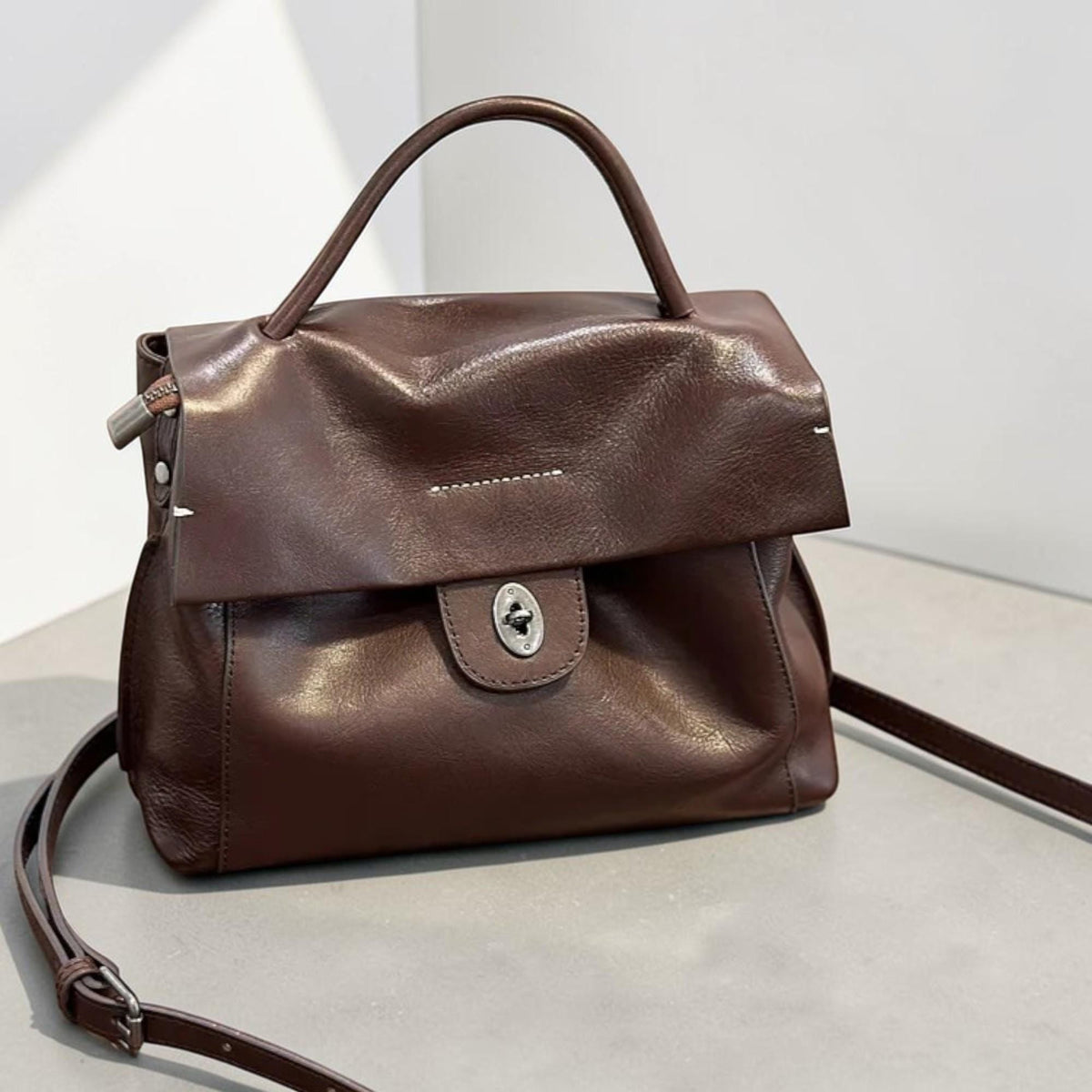 May include: A brown leather handbag with a top handle and a detachable shoulder strap. The bag features a flap closure with a silver-toned clasp. The leather has a slightly wrinkled texture, and the bag appears to be empty.