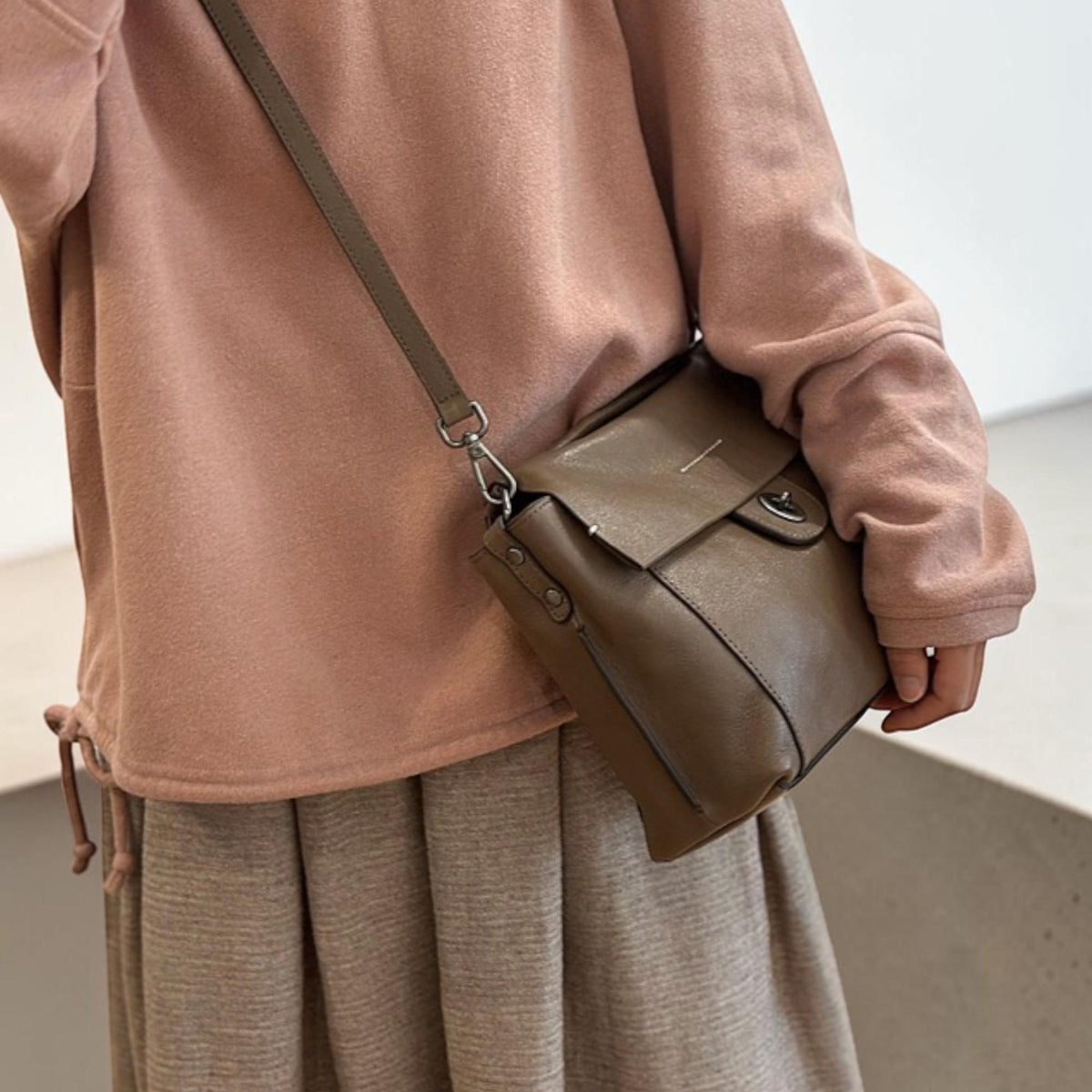 May include: A brown leather crossbody bag with a flap closure and a silver-tone clasp. The bag has a long, adjustable strap. The bag is shown against a pink sweater and a gray skirt.
