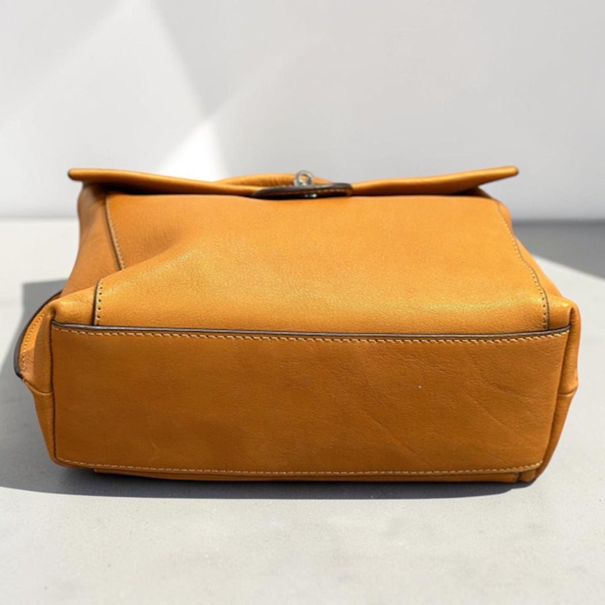 May include: A mustard-colored leather handbag with a flap closure. The bag has a rectangular shape with a stitched bottom and a metal clasp. The leather appears smooth and the bag is empty.