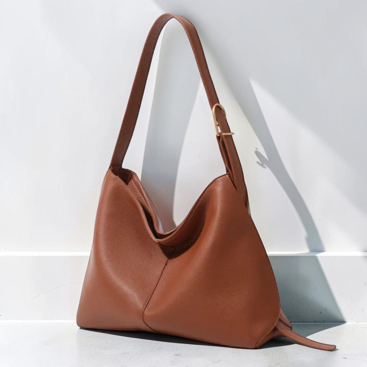 May include: A medium brown hobo-style shoulder bag with a smooth, matte finish. The bag features a single wide strap with a gold-toned metal buckle for length adjustment, a slouchy silhouette, and a central vertical seam detail on the front panel.