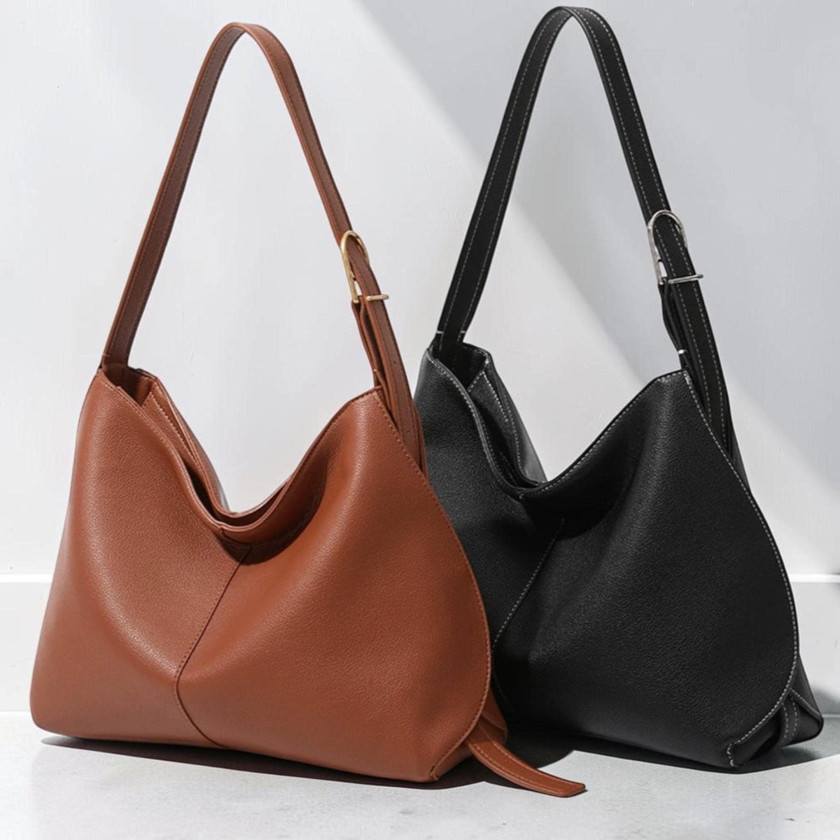 May include: Two hobo-style shoulder bags, one in a solid tan color and the other in black. Each bag features a single adjustable strap with a metal buckle, a smooth exterior finish, and a distinct diagonal seam detail across the front panel.
