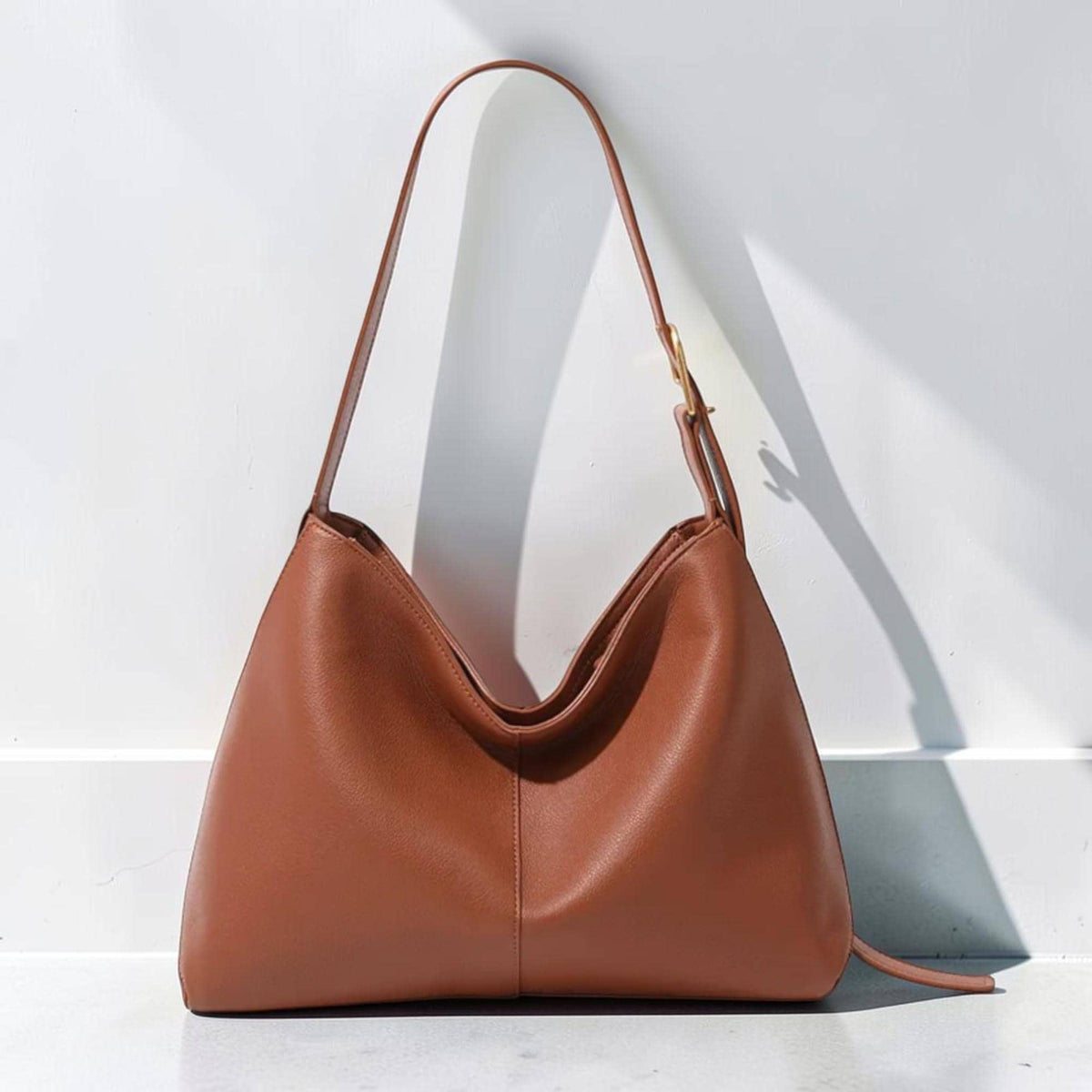 May include: A medium-sized, smooth, cognac-brown shoulder bag with a slouchy, trapezoidal silhouette. The bag features a single adjustable strap with a gold-toned metal buckle, a central vertical seam, and a wide, open-top main compartment.