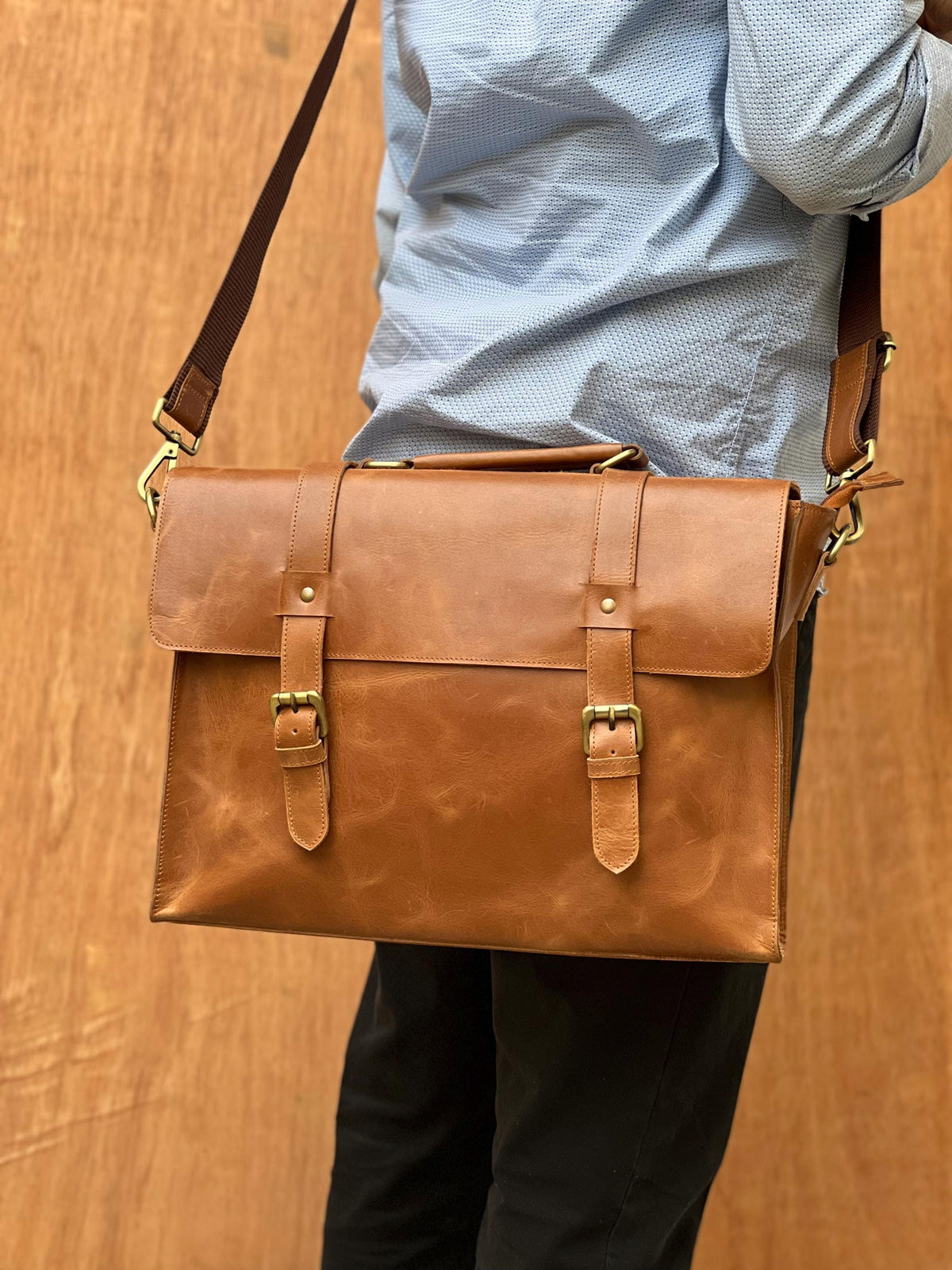 May include: A brown leather briefcase with a shoulder strap. The bag has two buckle closures and a top handle. The leather has a natural grain and a warm, reddish-brown color. The hardware is gold-colored.