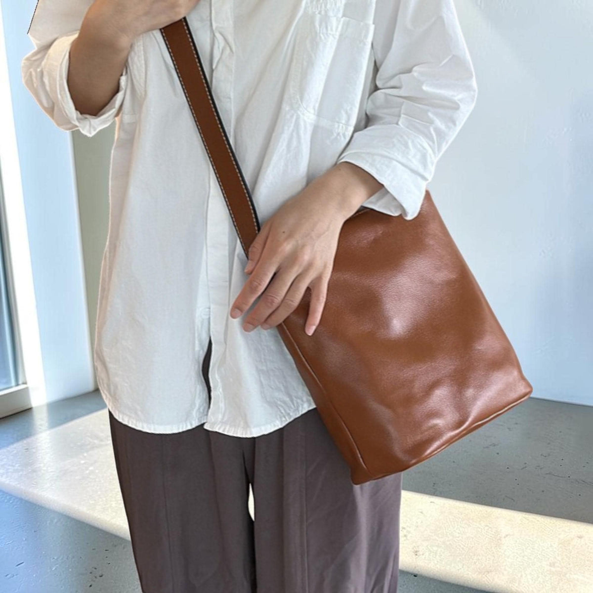 May include: A brown leather shoulder bag with a long strap. The bag is rectangular with rounded corners. The strap is brown with white stitching. The person is wearing a white shirt and brown pants.