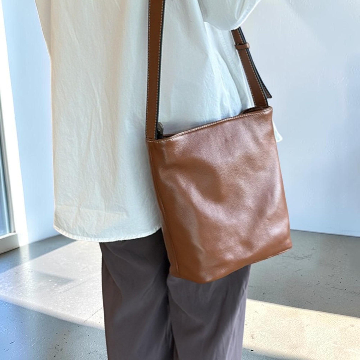 May include: A brown leather crossbody bag with a long strap. The bag is rectangular with a smooth surface. The strap is attached to the top corners of the bag. The bag is worn over the shoulder.
