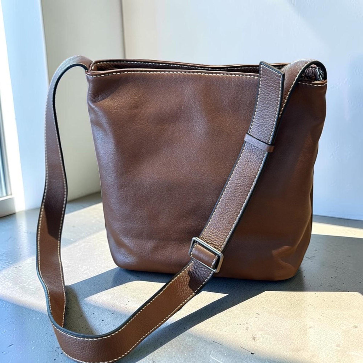 May include: A brown leather crossbody bag with a long adjustable strap. The bag has white stitching along the seams and a silver-colored buckle. The bag is a bucket style and appears to be empty.