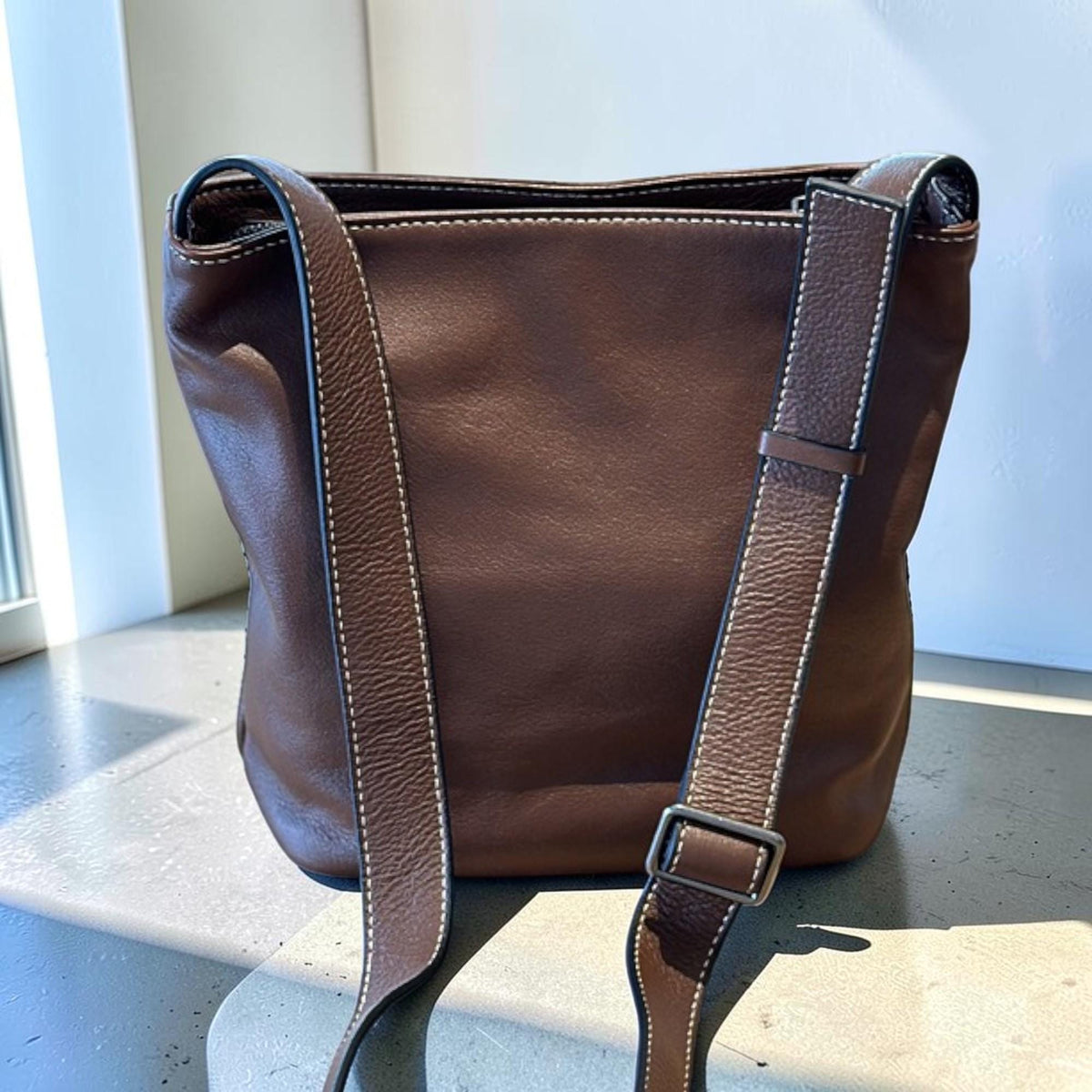 May include: A brown leather crossbody bag with a rectangular shape and a long adjustable strap. The bag features white stitching along the edges and a silver-colored buckle. The bag is made of a smooth, durable leather.