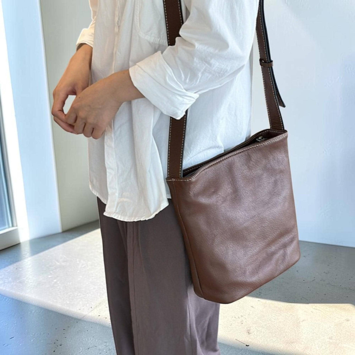 May include: A brown leather crossbody bag with a long strap. The bag has a simple, rectangular shape with a zipper closure. The strap is dark brown with white stitching. The bag is worn over the shoulder.