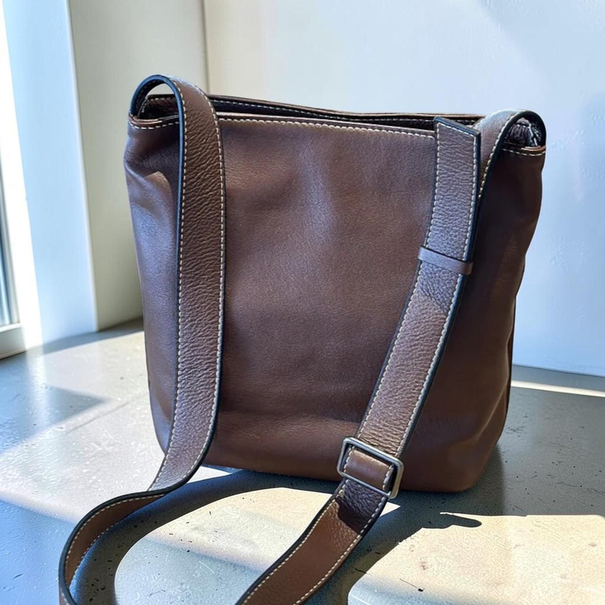 May include: A brown leather shoulder bag with a rectangular shape. The bag has a long, adjustable strap and white stitching details. The bag is made of a soft, supple leather.