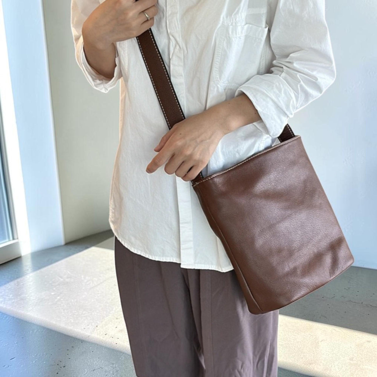 May include: A brown leather crossbody bag with a rectangular shape. The bag has a long strap with white stitching and a simple design. The bag is worn over the shoulder.
