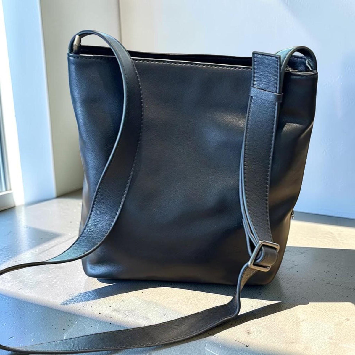 May include: A black leather shoulder bag with a rectangular shape. The bag has a long, adjustable strap and a silver-colored buckle. The bag is sitting on a light-colored surface.