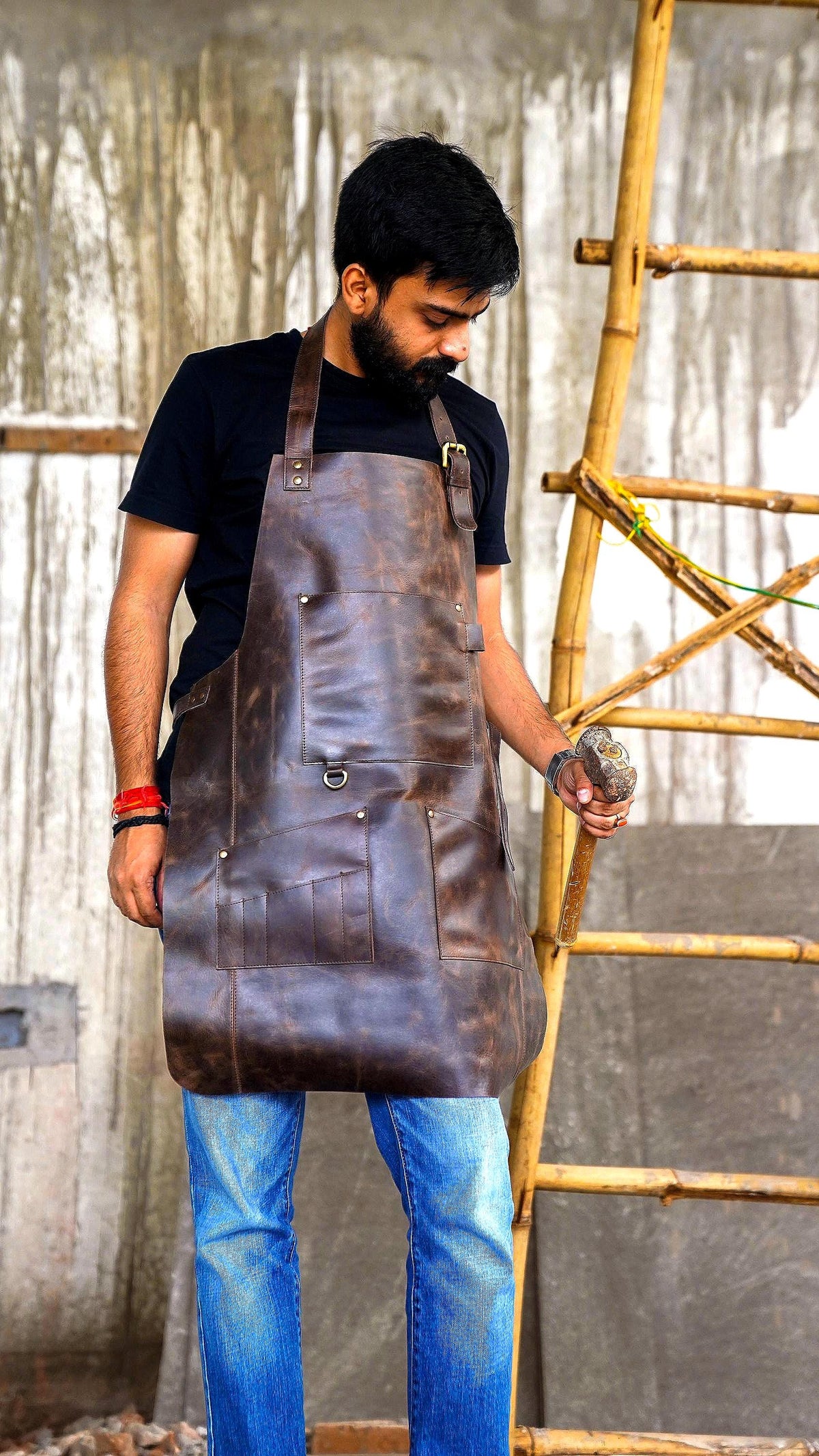 May include: A dark brown leather apron featuring a neck strap with a metal buckle, a large chest pocket, and multiple lower utility pockets with metal rivets. The apron includes a small metal ring attachment and visible stitching along the edges.