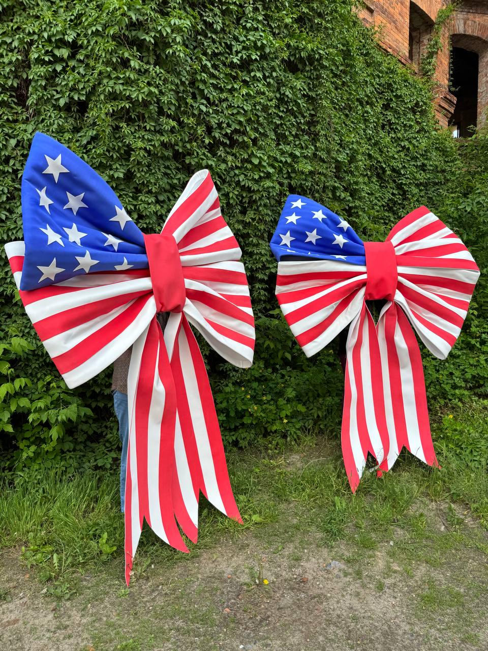May include: Two large decorative bows with a patriotic American flag design. The bows feature a blue section with white stars and red and white stripes. The bows are made of a sturdy material and have long, flowing tails.