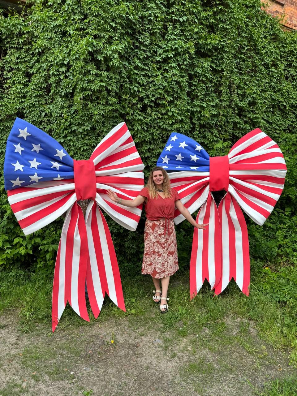 May include: Two large decorative bows with a patriotic American flag design. The bows feature a blue section with white stars and red and white stripes. The bows are displayed outdoors against a backdrop of green foliage.