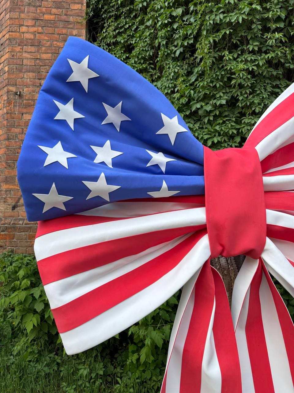 May include: A large decorative bow featuring the American flag design. The bow has a blue section with white stars, and red and white stripes. The bow is tied with a red band in the center. The bow is likely for outdoor decoration.