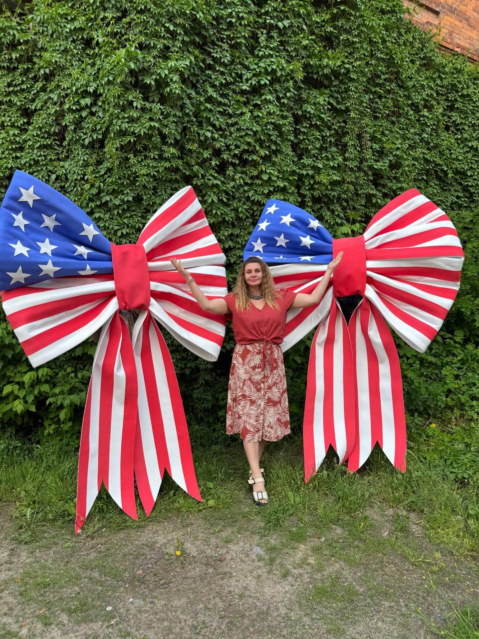 May include: Two large decorative bows with a patriotic American flag design. The bows feature blue sections with white stars and red and white striped ribbons. A person stands between the bows in a red top and patterned skirt.