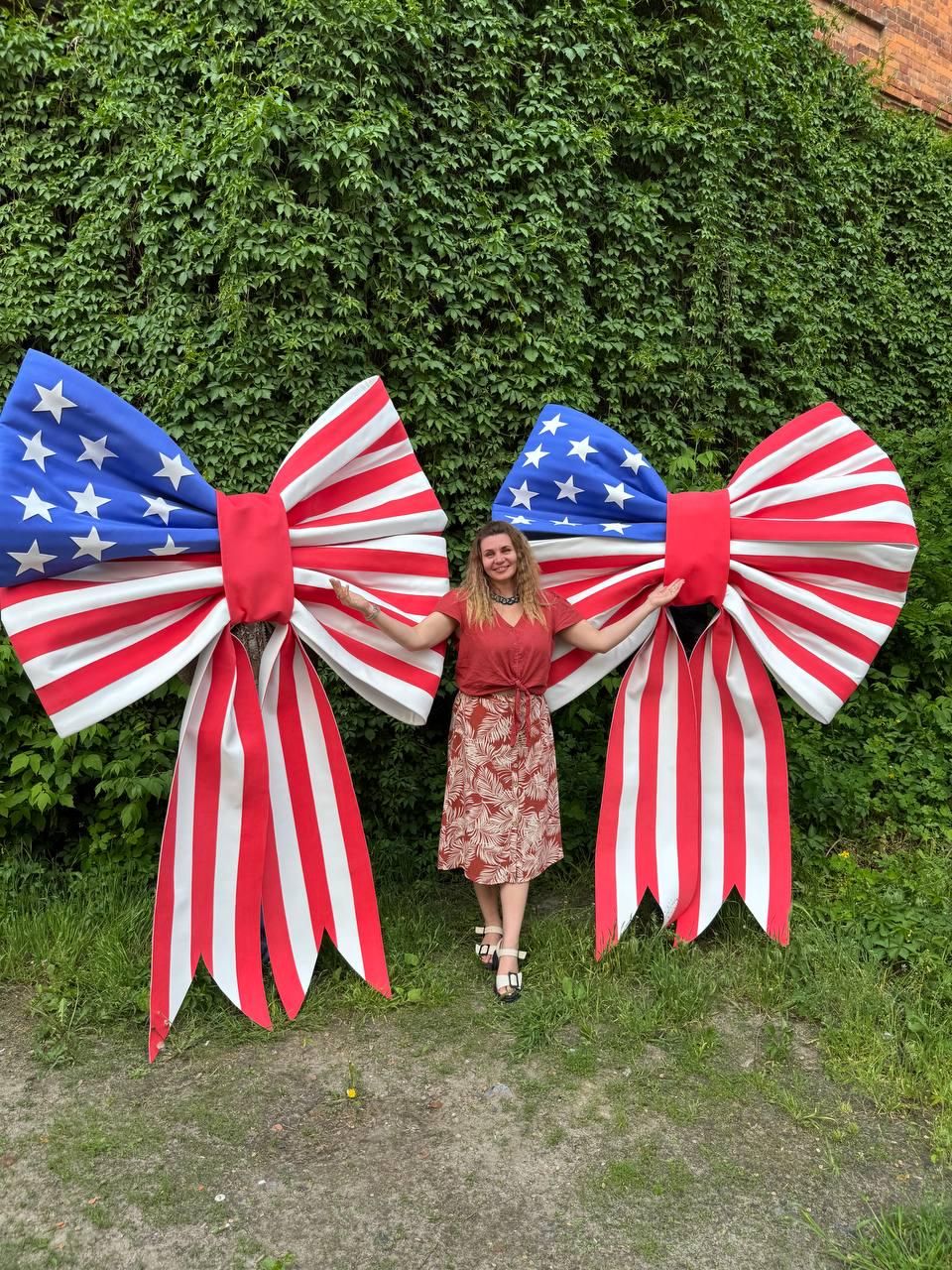 May include: Two large decorative bows in the style of the American flag. Each bow features a blue section with white stars and red and white striped ribbons. The bows are displayed outdoors against a backdrop of green foliage.