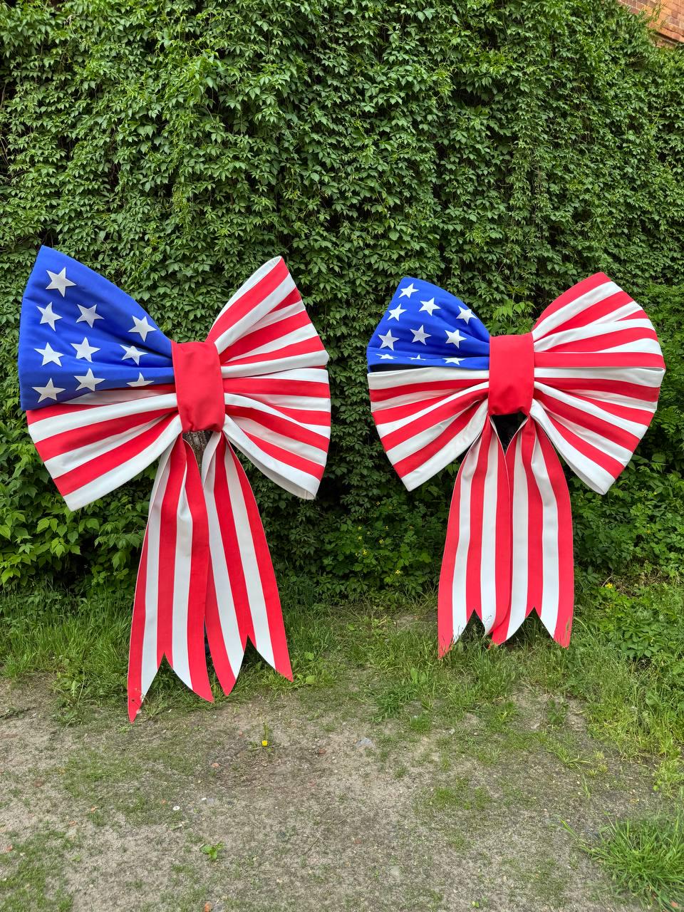 May include: Two large decorative bows with a patriotic theme. The bows feature a blue section with white stars, red and white stripes, and long red and white striped tails. They are set against a backdrop of green foliage.