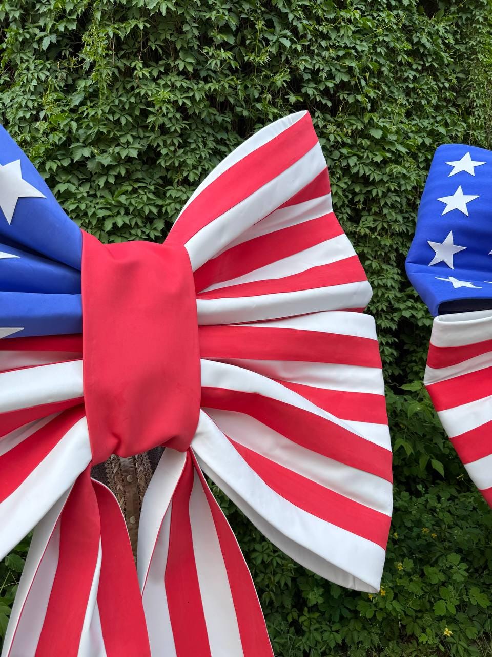 May include: Large patriotic bow decorations in red, white, and blue. The bows feature red and white stripes and blue sections with white stars, resembling the American flag. The bows are set against a backdrop of green foliage.