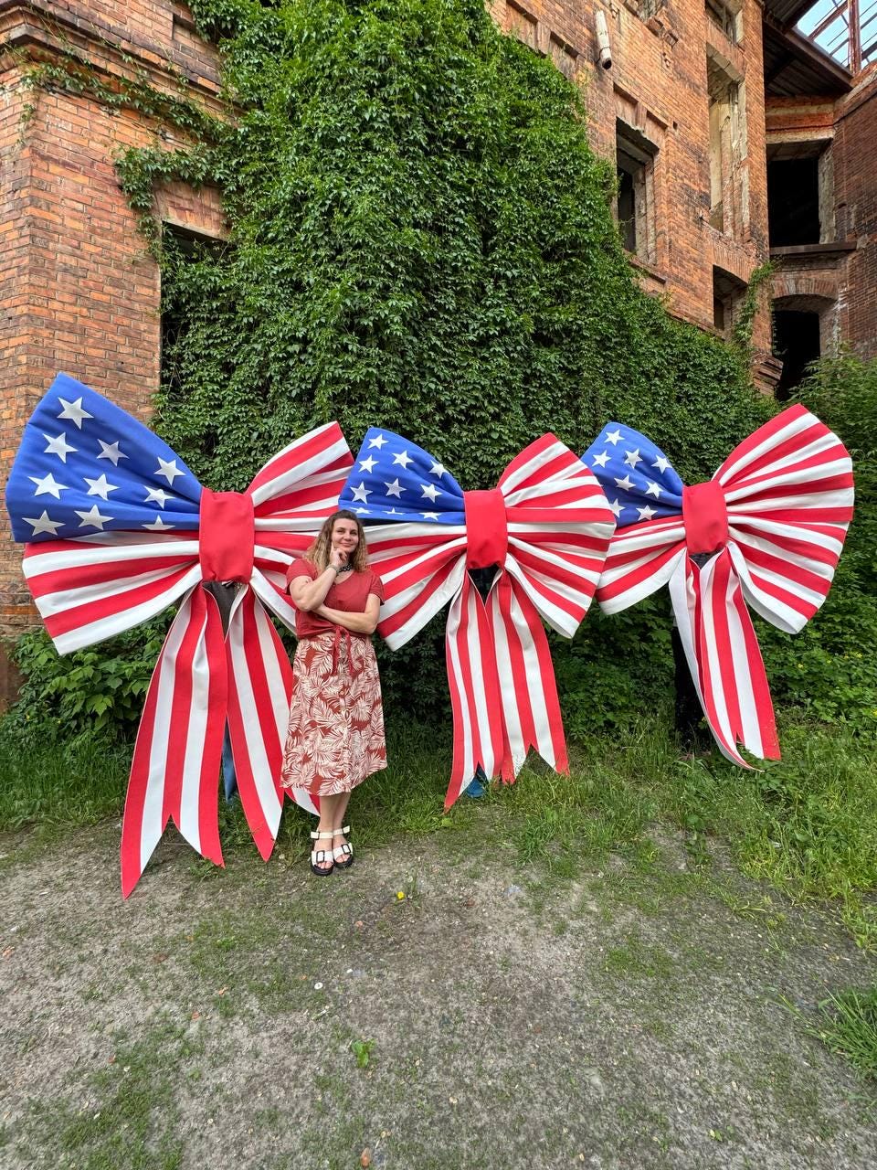 May include: Three large decorative bows in red, white, and blue, resembling the American flag. The bows have a blue section with white stars and red and white striped ribbons. The bows are displayed outdoors in front of a brick building.
