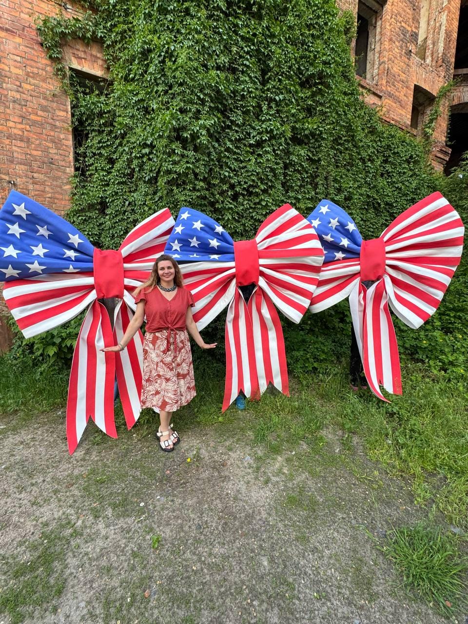 May include: Three large patriotic bows with red and white stripes and blue sections with white stars. The bows are displayed outdoors with a person standing in front of them. The background includes green foliage and a brick building.