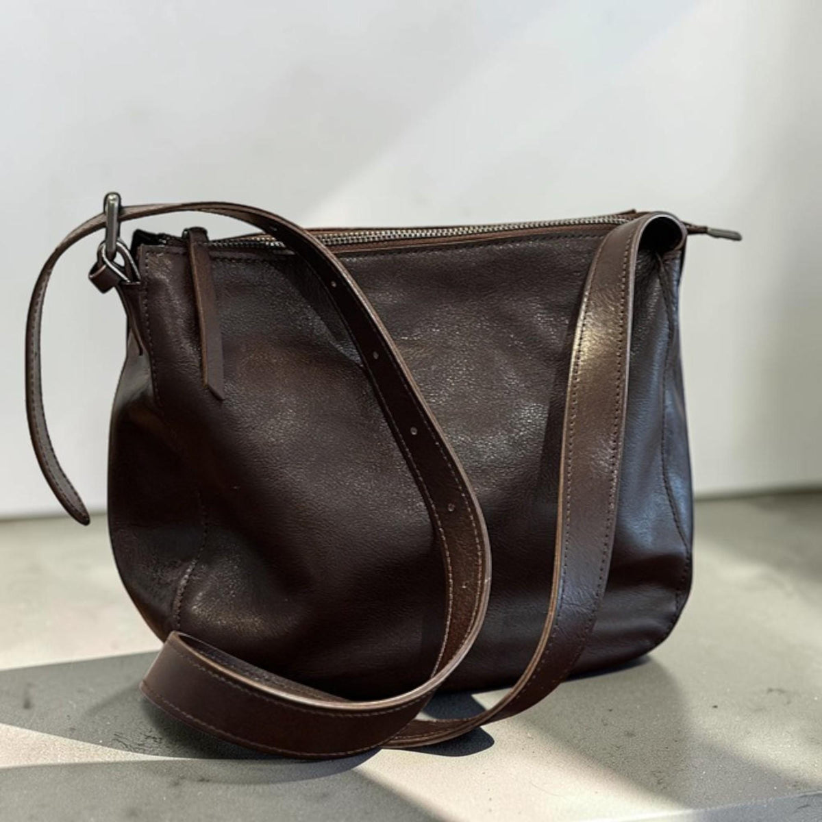 May include: A dark brown leather shoulder bag with a long adjustable strap. The bag has a zipper closure and a rounded shape. The leather appears to be textured and has a slight sheen. The bag is sitting on a light gray surface.