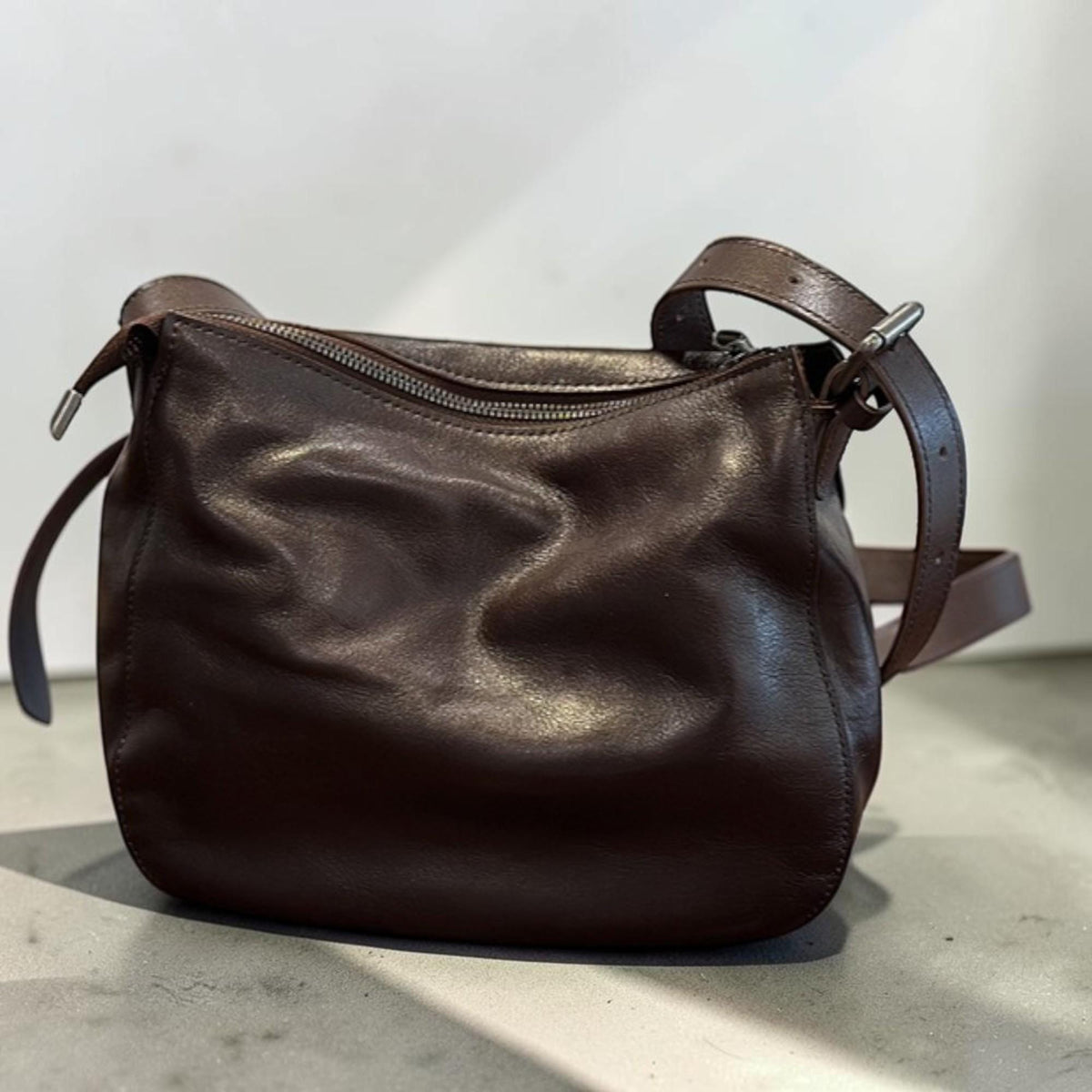 May include: A dark brown leather shoulder bag with a curved shape. The bag has a zipper closure and a long, adjustable strap. The leather appears soft and has a slightly wrinkled texture. The bag is designed for everyday use.