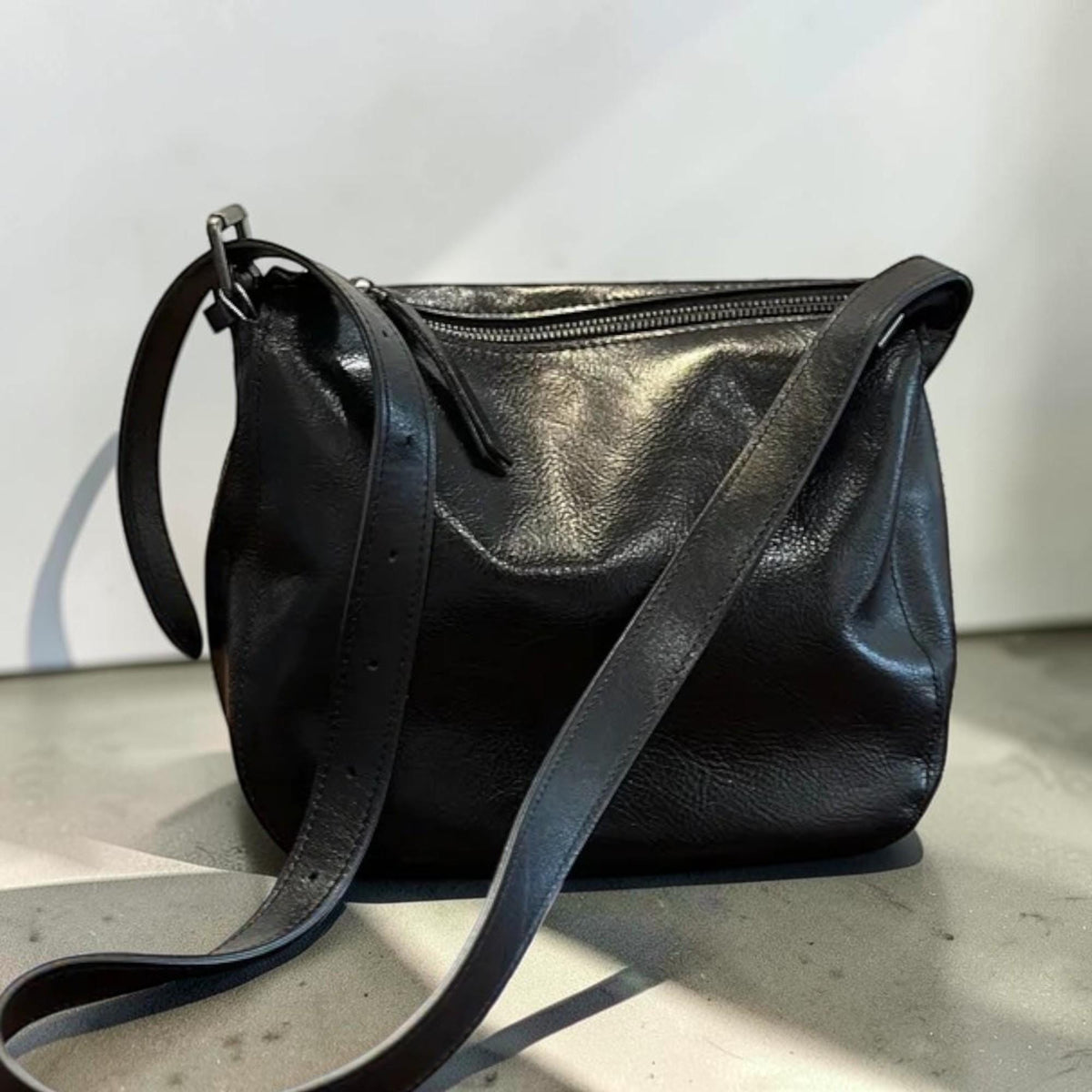 May include: Black leather crossbody bag with a long adjustable strap. The bag has a zippered top closure and a rounded shape. The leather appears to have a slightly textured finish, and the strap is attached with silver-toned hardware.