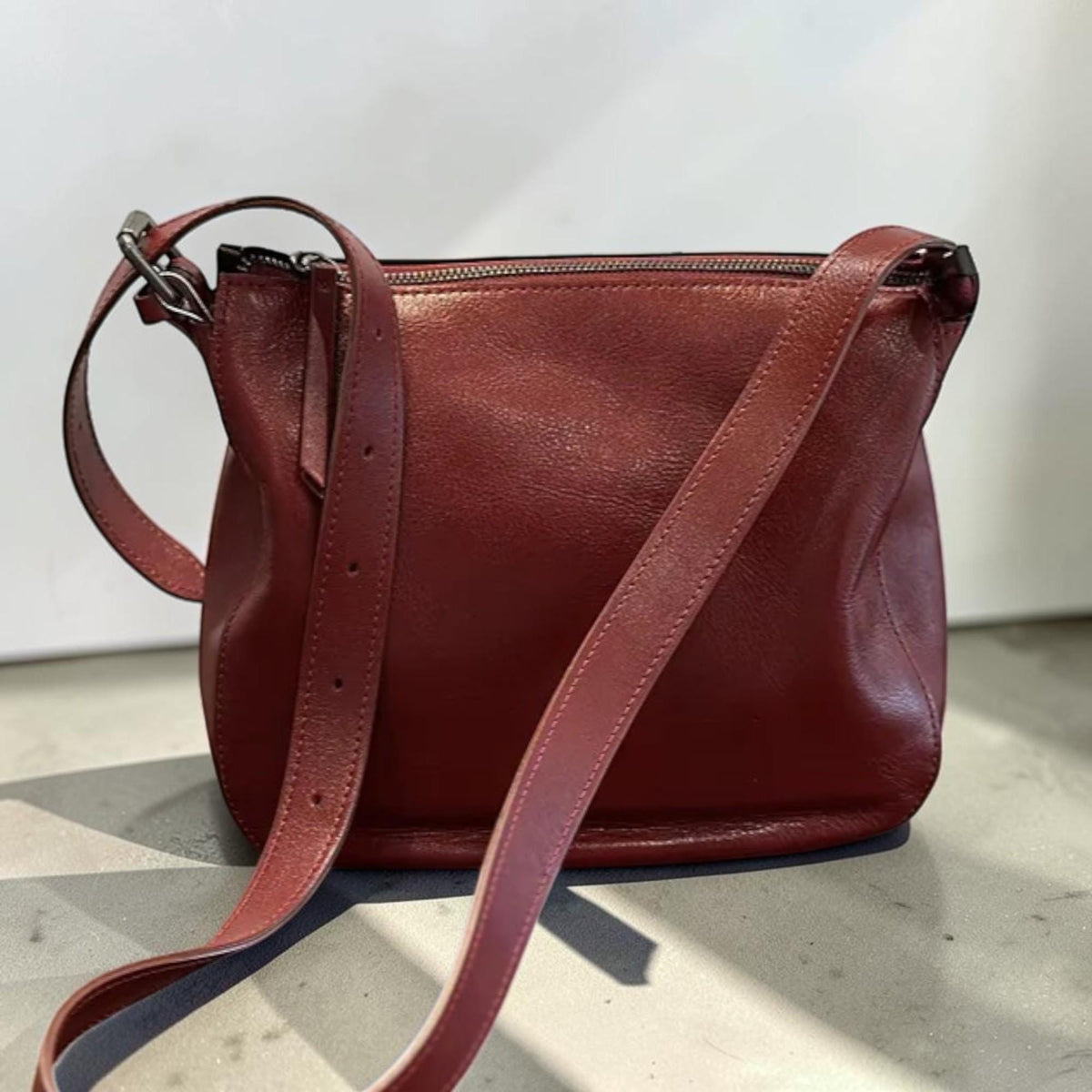 May include: A burgundy leather crossbody bag with a long, adjustable strap. The bag has a zippered top closure and a rounded shape. The strap is attached to the bag with silver-colored metal hardware.