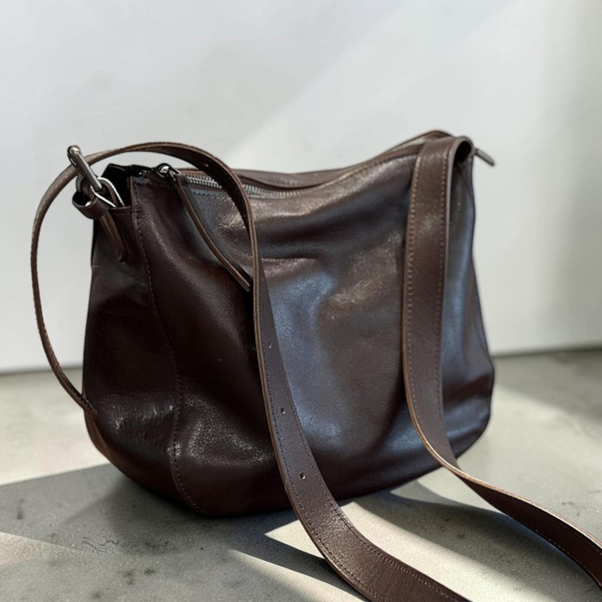 May include: A dark brown leather shoulder bag with a long adjustable strap. The bag has a zippered top closure and a rounded shape. The leather appears soft and slightly textured, with a subtle sheen. The bag is designed for everyday use.