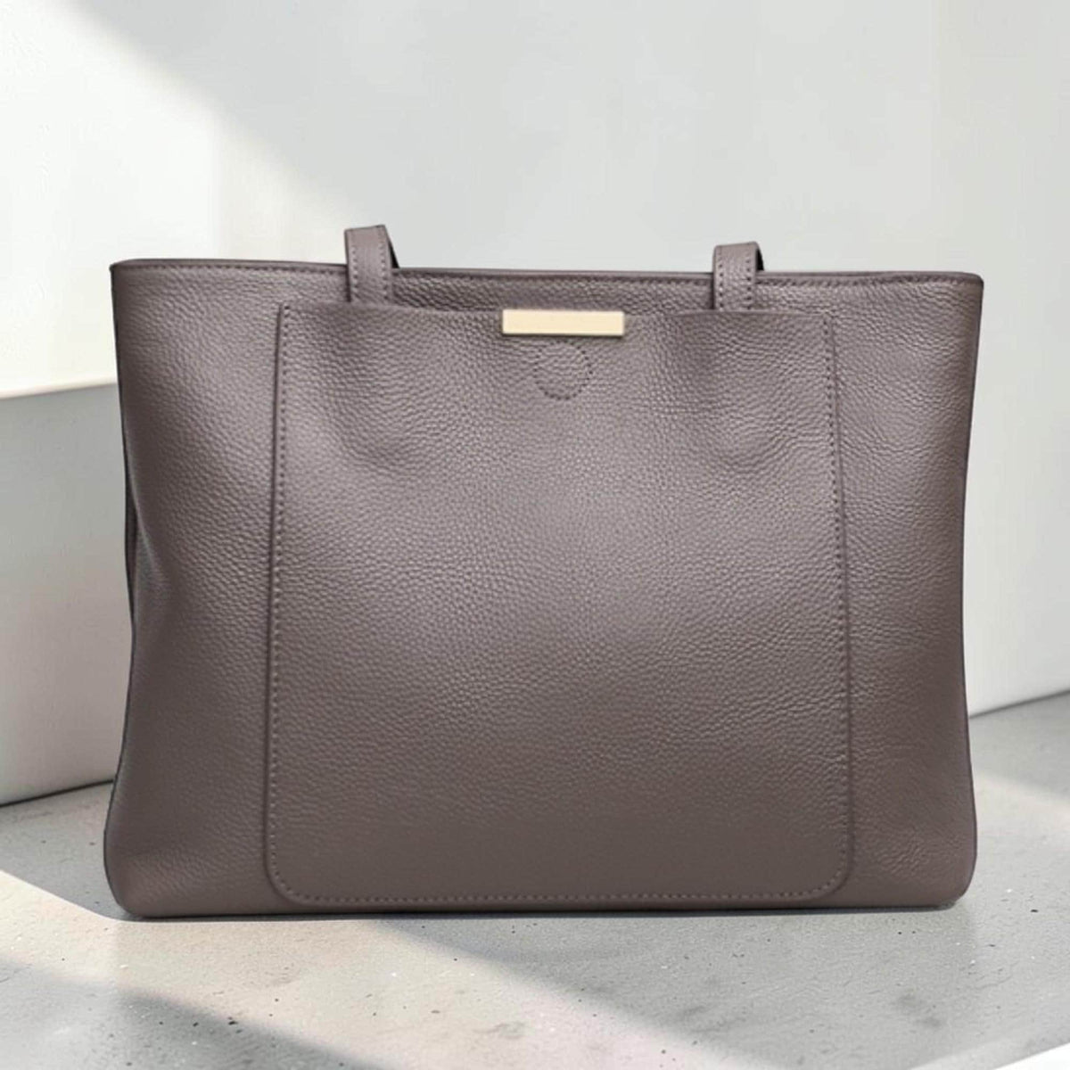 May include: A taupe-colored leather tote bag with a pebbled texture, featuring two top handles and a rectangular front pocket. The bag is accented with a horizontal gold-toned metal bar centered at the top edge of the front pocket, providing a clean look.