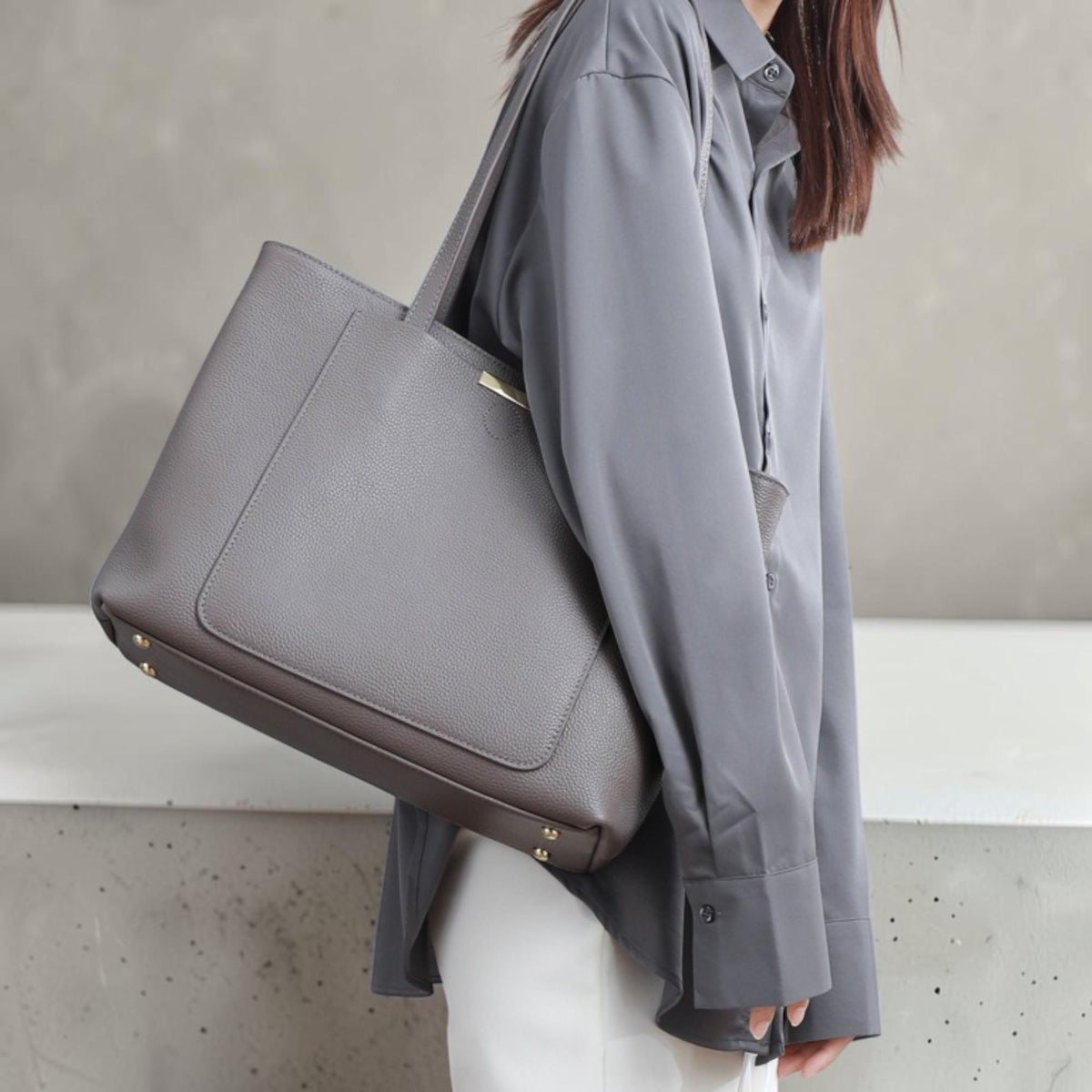 May include: A large, structured grey tote bag with a pebbled texture, featuring a rectangular front pocket, thin shoulder straps, and small gold-toned metal feet on the bottom base. The bag includes a thin gold-toned metal accent bar at the top.
