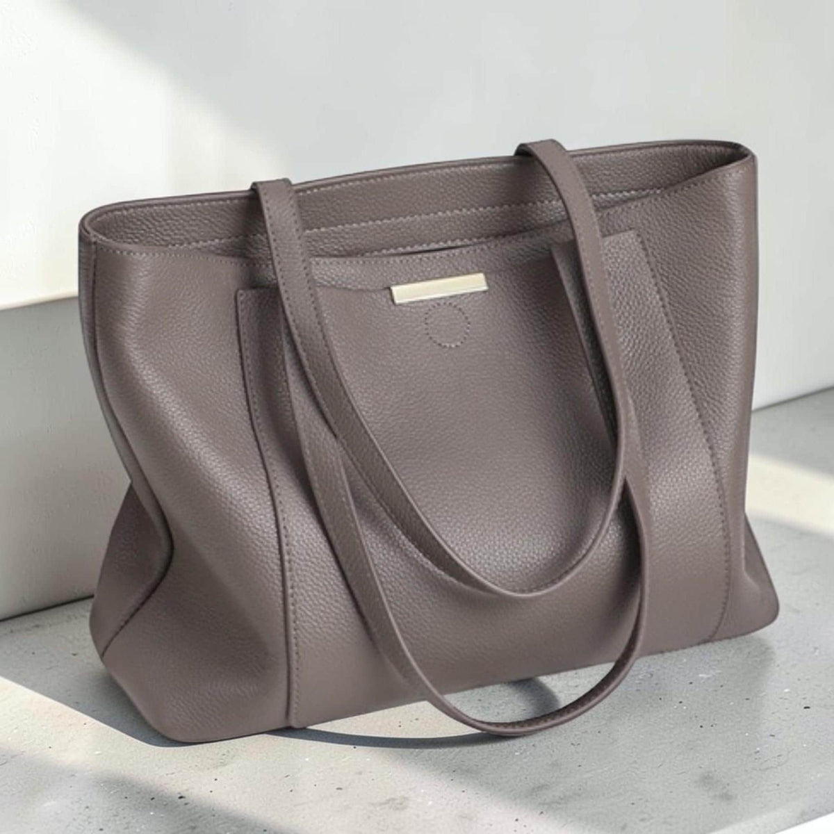 May include: A taupe-colored tote bag with a pebbled texture, featuring two long shoulder straps, a rectangular silhouette, and a small horizontal metallic bar accent centered on the upper front panel above a circular stitched detail.