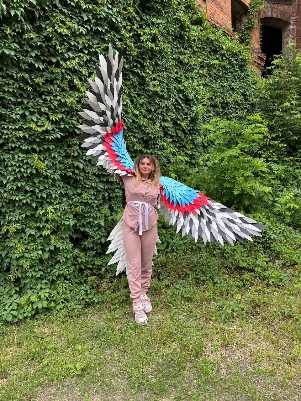 May include: Large wearable wings featuring layered feathers in shades of white, gray, black, red, and blue. The wings are attached to a dusty pink long-sleeved button-down shirt and matching pants, secured at the waist with a white ribbon tie.