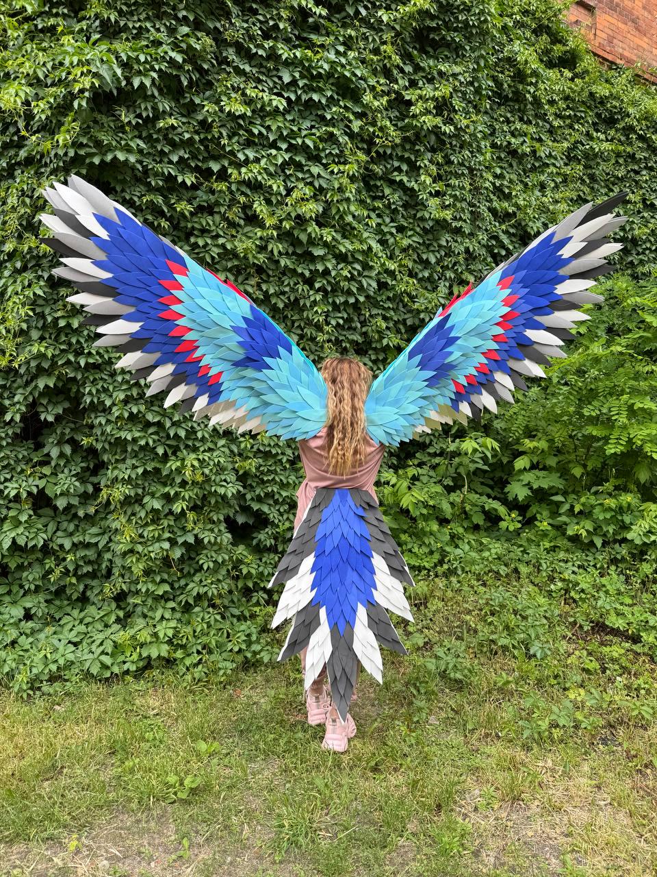 May include: Large wearable bird wings and a matching tail featuring layered feathers in shades of blue, light blue, white, gray, and red. The wingspan is wide and arched, while the tail consists of pointed, overlapping feathers in a downward pattern.
