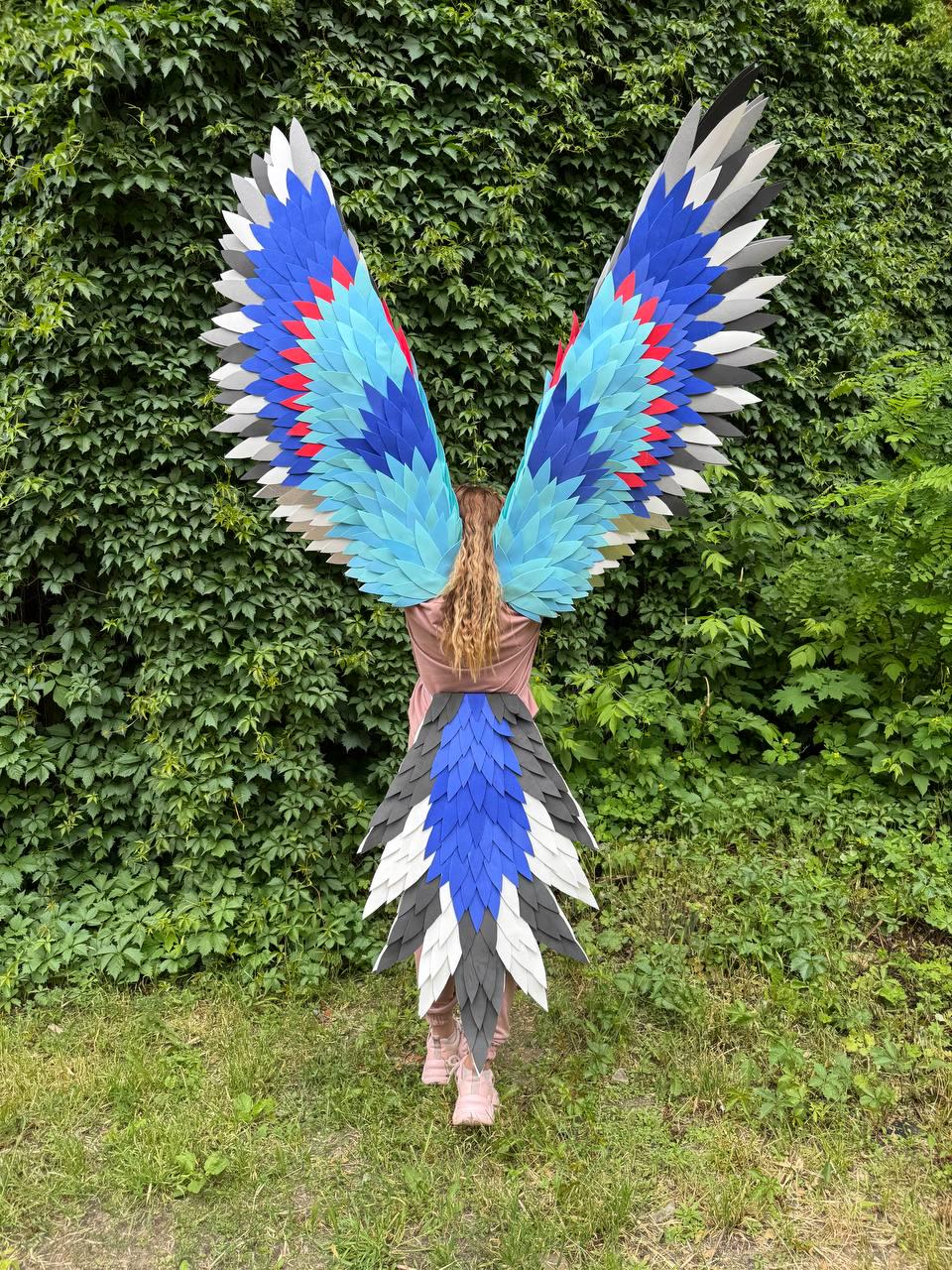 May include: Large wearable bird wings and a matching tail attachment featuring layered feathers in shades of royal blue, light blue, white, dark gray, and accents of bright red. The wings are extended upward and the tail hangs downward.