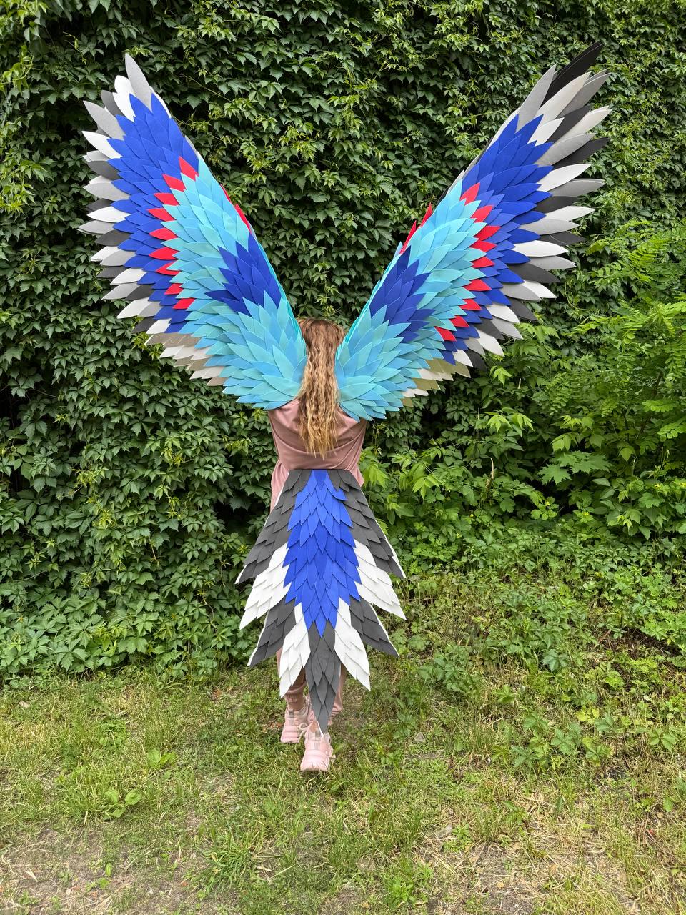 May include: Large wearable bird wings and a matching tail featuring layered feathers in shades of royal blue, light blue, white, gray, and accents of red. The wings are extended upward and outward, while the tail hangs down from the waist area.