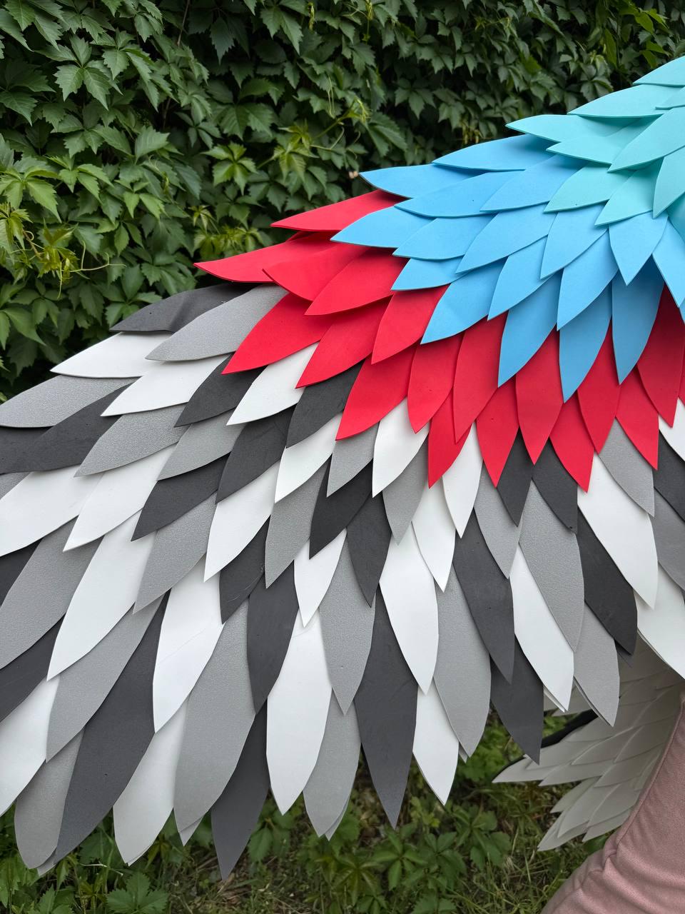 May include: A pair of large decorative wings constructed from layered, leaf-shaped foam pieces. The feathers are arranged in a gradient pattern featuring bright blue, vibrant red, dark gray, light gray, and white colors in a textured design.