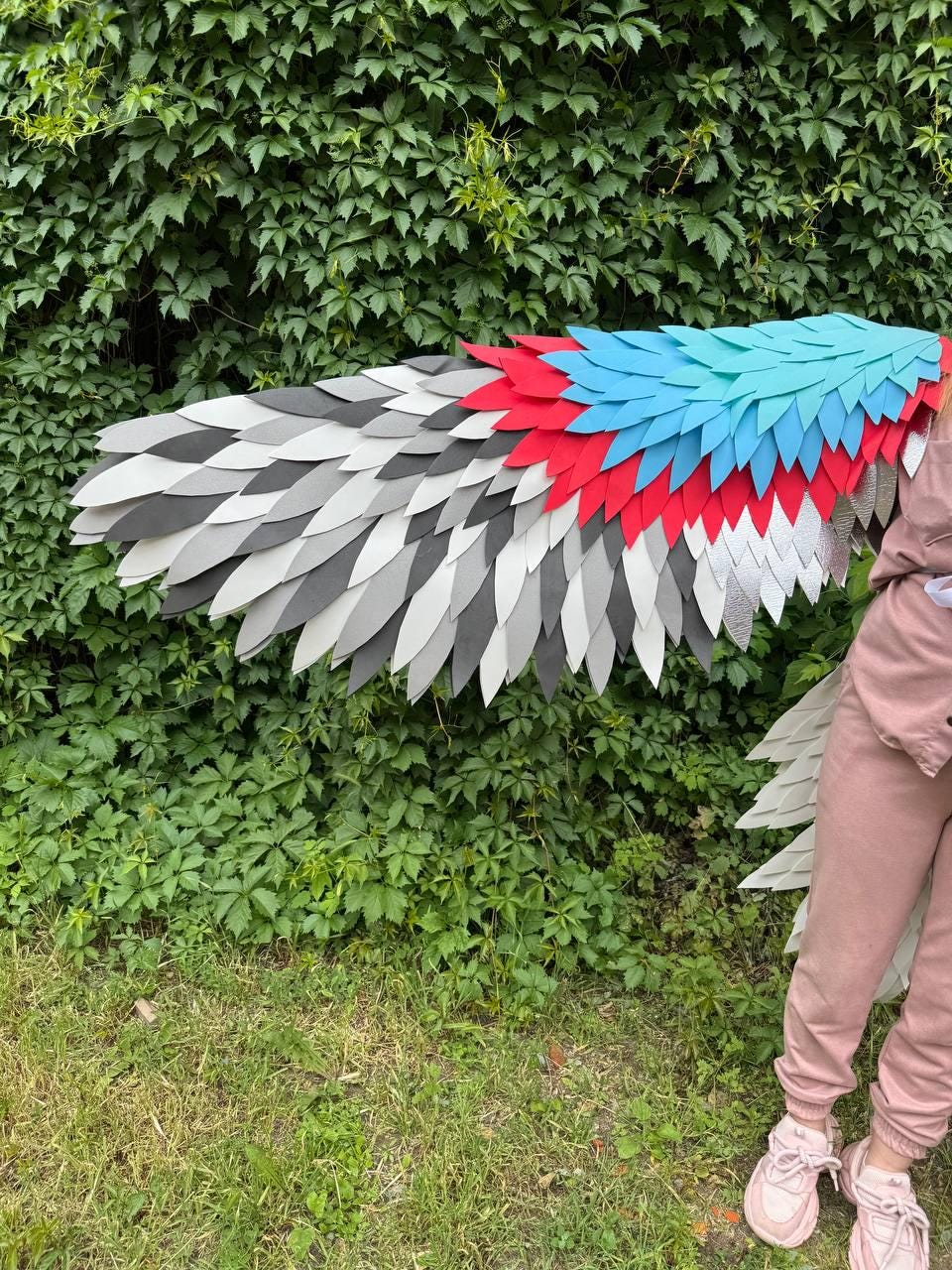 May include: A large set of costume wings featuring layered, feather-shaped pieces in a gradient of colors. The wing displays a pattern of light blue, bright red, dark gray, light gray, and white segments arranged in rows to mimic bird plumage.