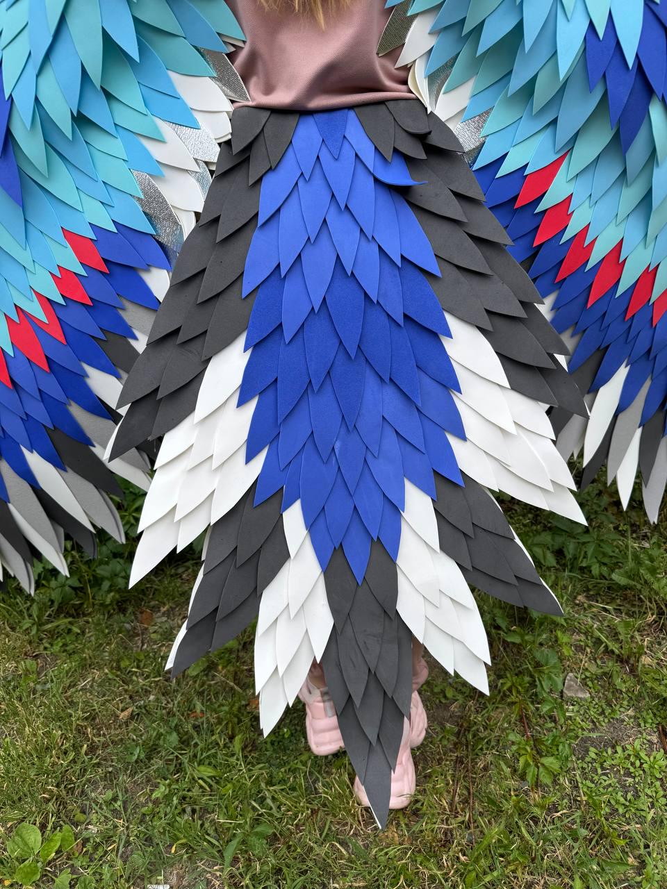 May include: A costume featuring large, layered wings and a tail made of pointed, feather-like shapes in shades of blue, black, white, and red. The wings extend outward, while the tail hangs down, creating a bird-like appearance with overlapping segments.