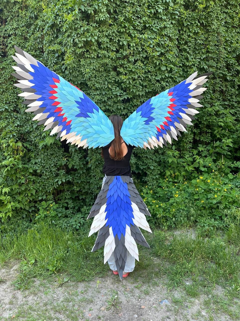May include: Large costume wings and a matching tail featuring layered, feather-shaped pieces in shades of bright blue, light blue, red, dark gray, and white. The wings extend outward, and the tail hangs downward, creating a bird-like appearance.
