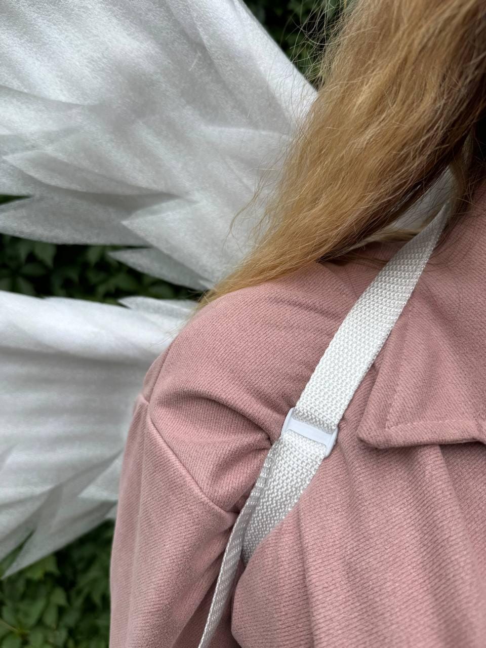 May include: A close-up shot of a light pink jacket with a white woven strap and a white plastic buckle. The strap is diagonally across the jacket. White fabric is visible in the background.