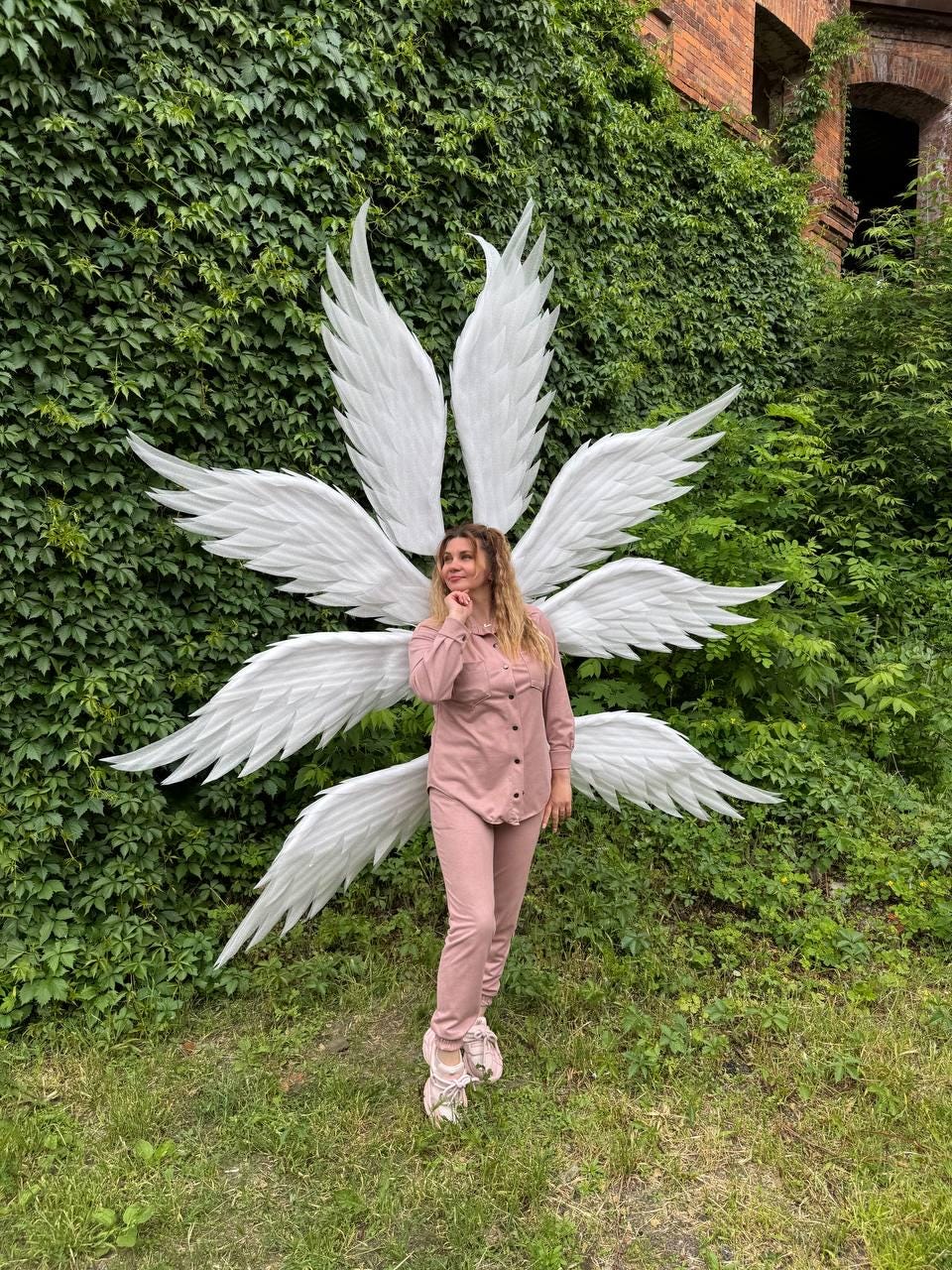 May include: Large, white angel wings with multiple layers of feathers are displayed outdoors. The wings are positioned behind a person wearing a pink outfit. The background features lush green foliage and a brick building.